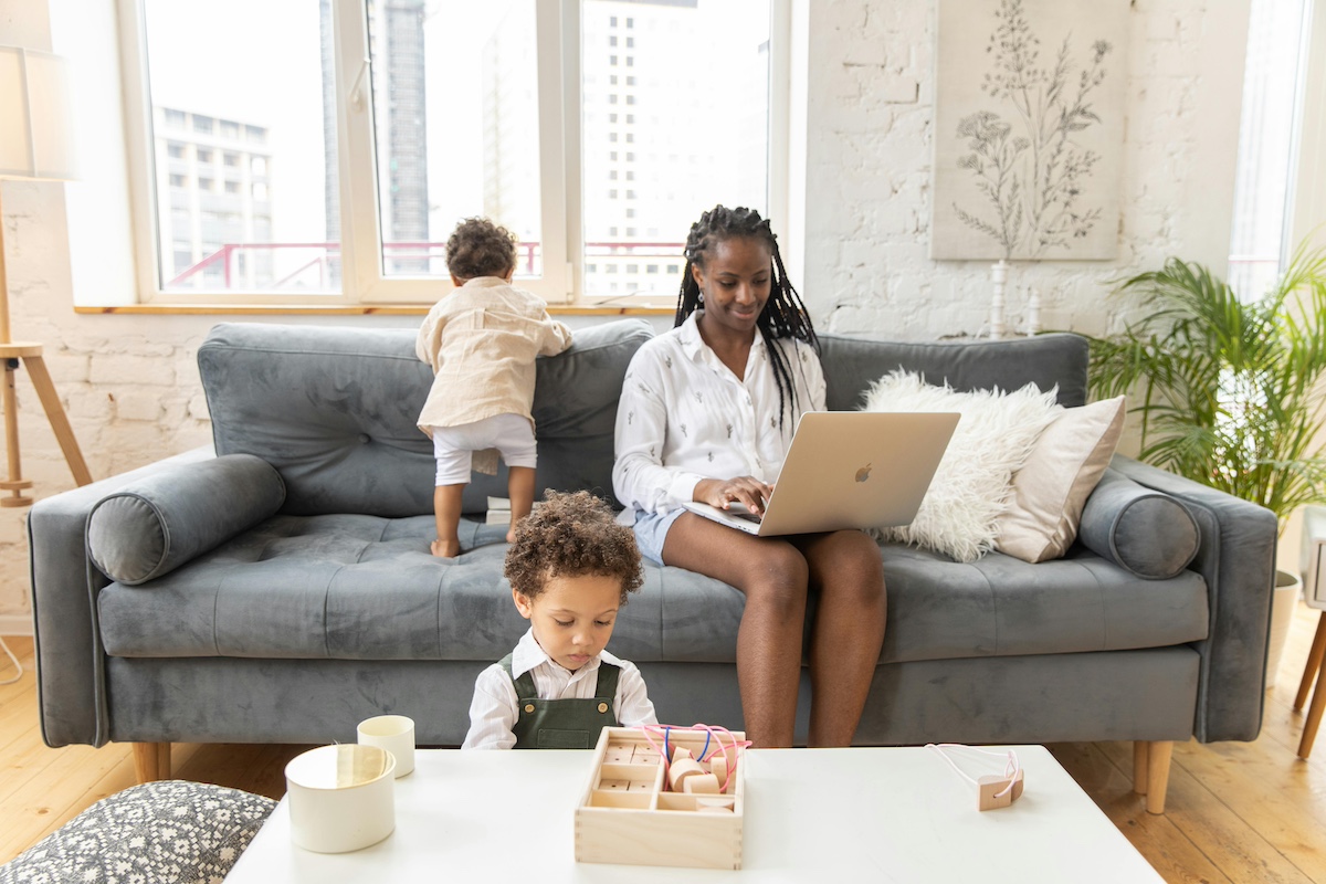 Woman sitting at laptop working on her couch next two her two children.